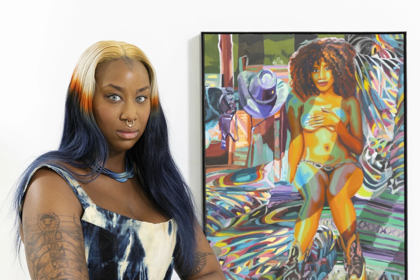 A female presenting person with brown skin poses next to a painting of a nude female covering their breasts.