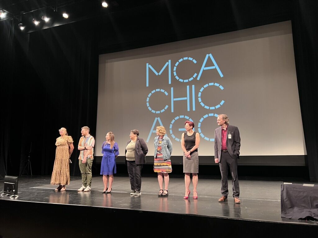 Seven people stand on a stage with an MCA Chicago logo projected on a screen behind them.