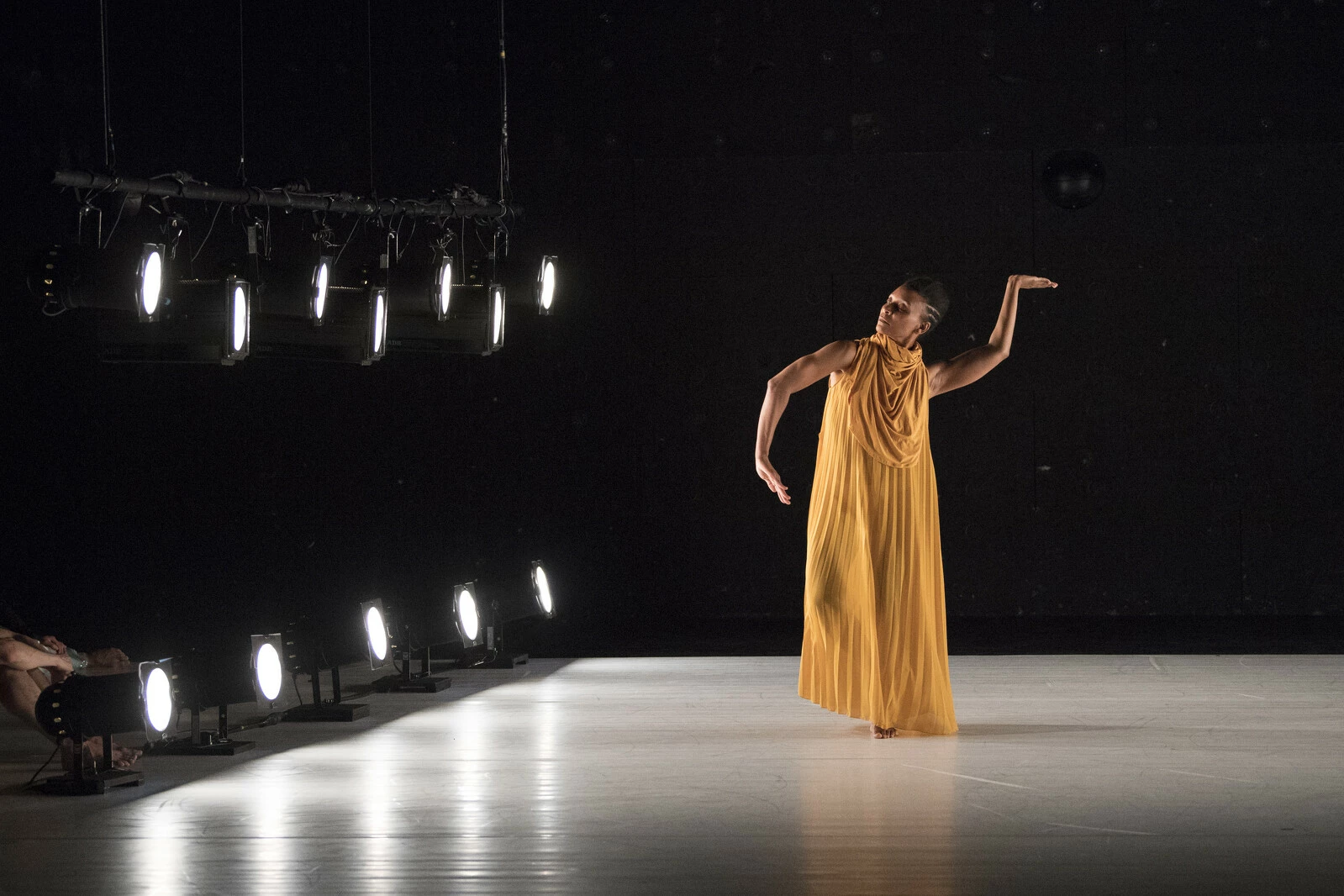 A dancer on a stage dressed in marigold yellow poses with their left arm raised into an S shape; their right arm in a C shape with their hand pointed to their hip.