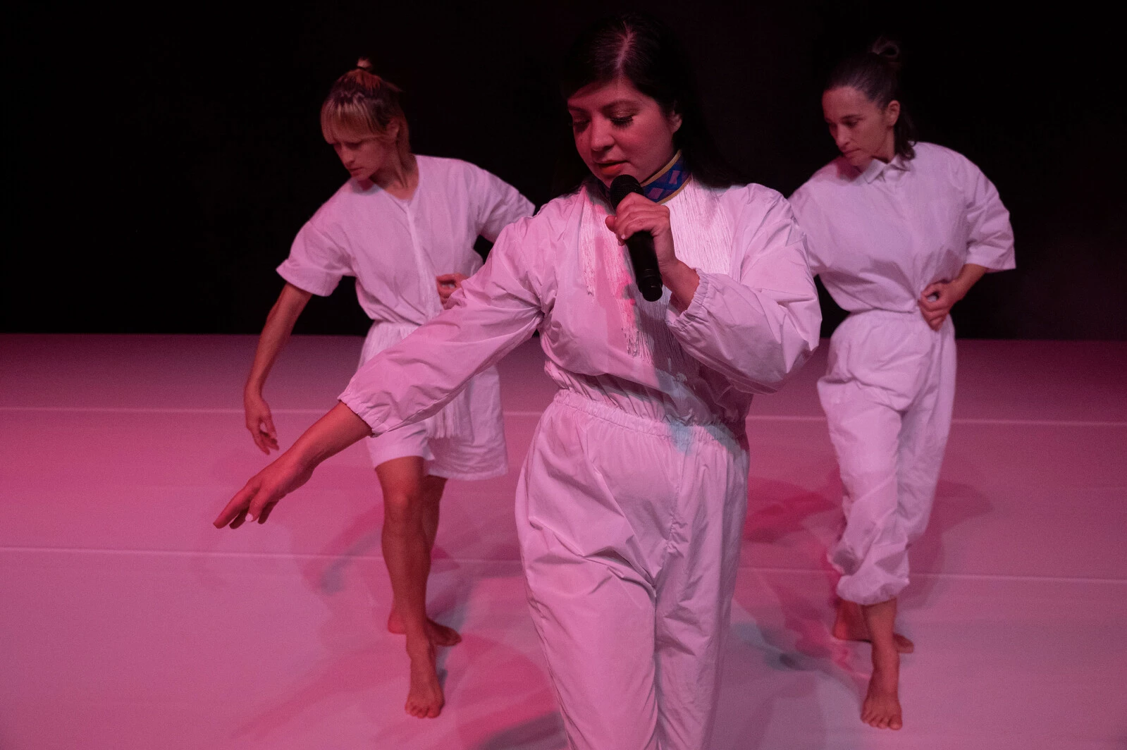 Three female-presenting people dressed in white performing on a stage lit with red light. The middle person in front holds a microphone.