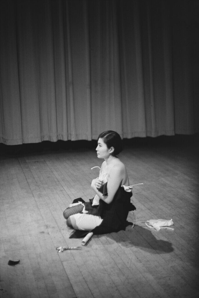 Archival photo of Yoko Ono sitting on the ground wearing scraps of clothing