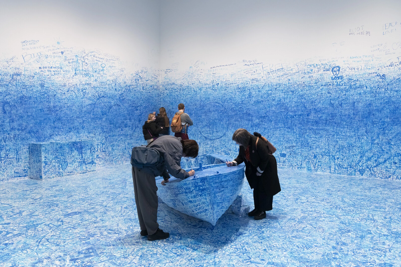 People standing over a blue boat in a white walled room covered in blue writing, including on the walls and floor surrounding it
