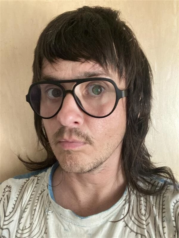 A person with long brown hair, thick rimmed glasses, and a short mustache posing against a beige wall