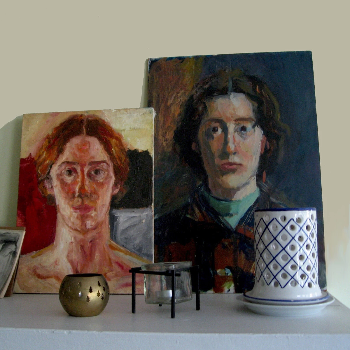 Two paintings of a female-presenting person painted in different styles lean against a beige wall with candles and other trinkets in front of them