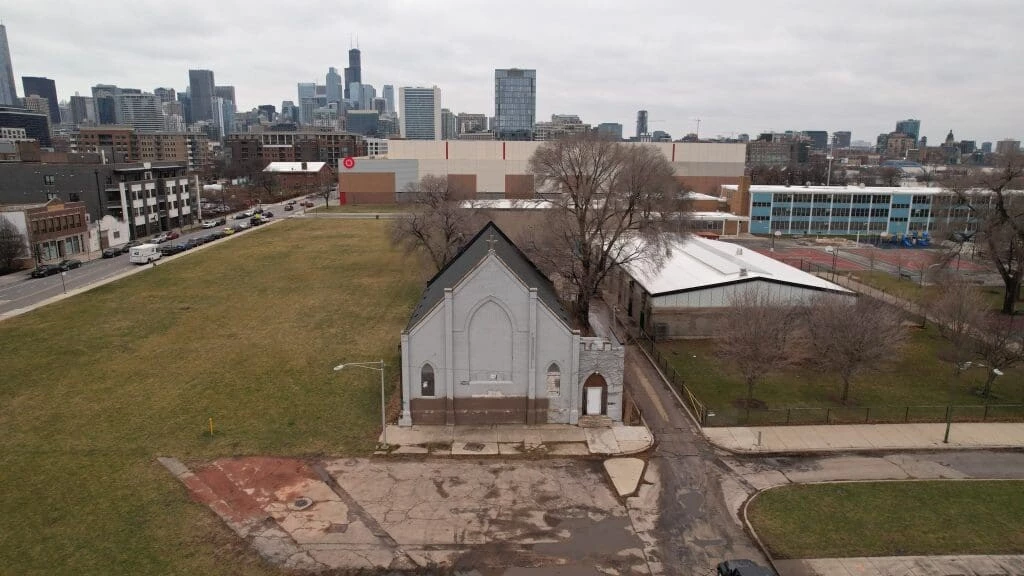 An old grey church with bricked-up windows among several empty lots and a warehouse. In the background is a Target store; a cityscape lies beyond that.