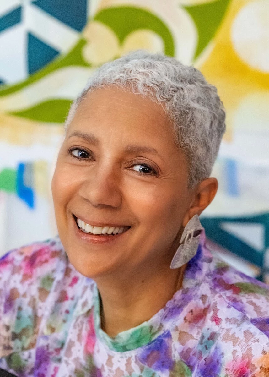 Portrait of a smiling person with short gray hair, wearing a colorful tie-dye shirt and leaf-shaped earrings, with a vibrant abstract mural in the background.