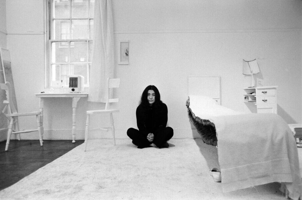 Black and white photo of Yoko Ono sitting crossed legged in a room where everything, from the bed to the artwork hanging on the wall, only has half