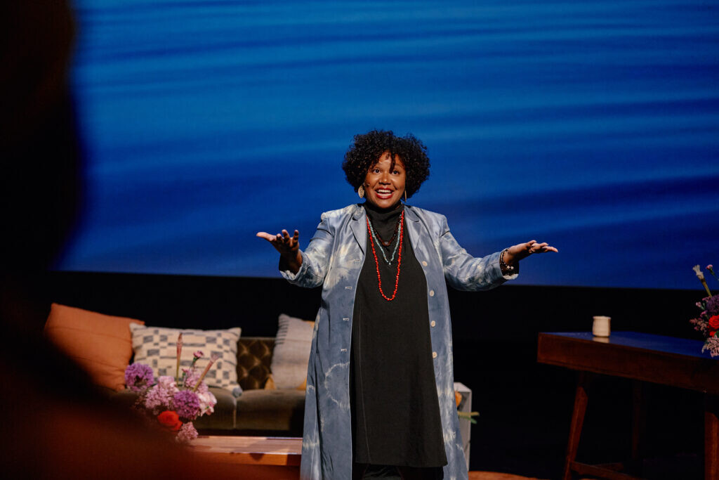 A female-presenting person offers their hands to an unseen audience on a stage with a pillow-filled couch and a blue, water-like projection behind them.