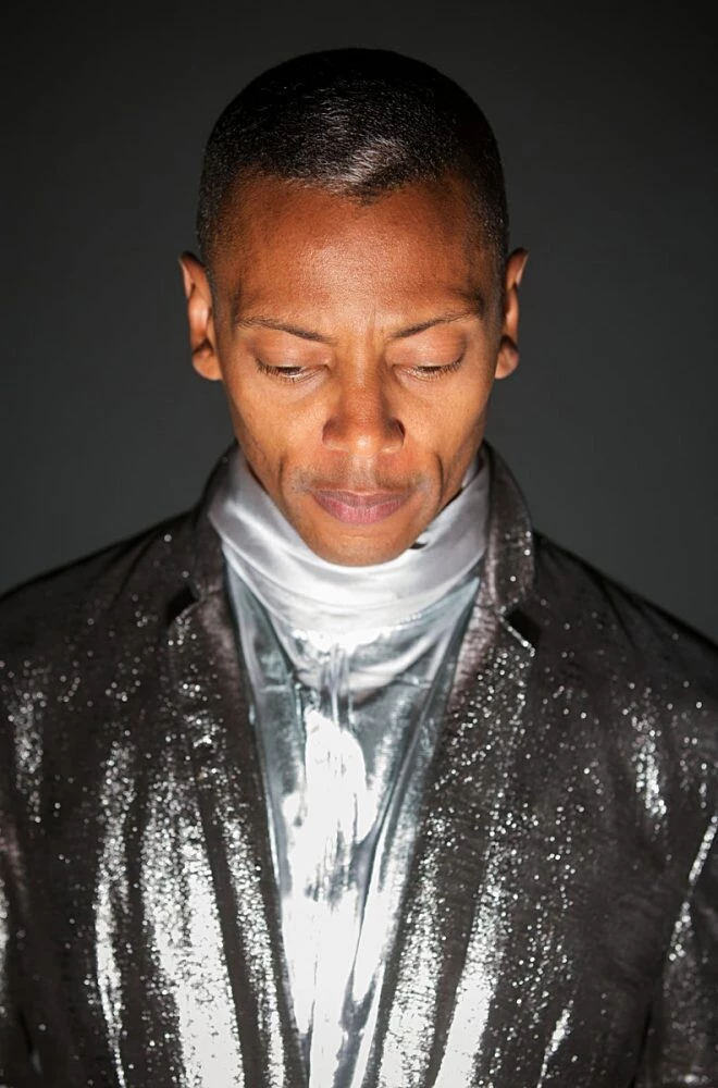 A person dressed in a shiny silver suit looks down at a light shining from below to create a dramatic effect