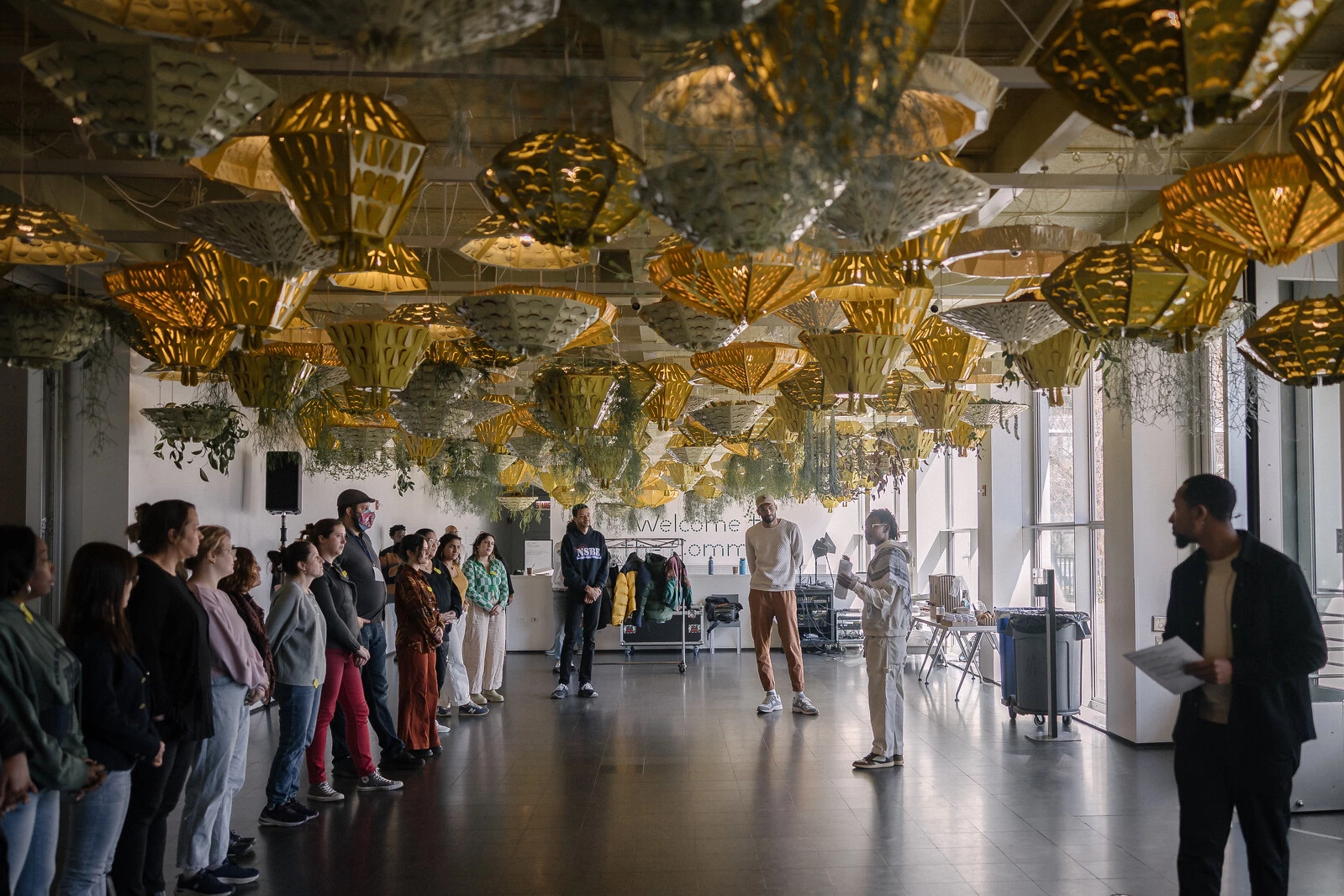A line of adults face two people on the other side of a room filled with hanging planters