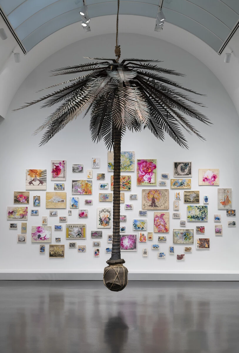 A palm tree hangs from the ceiling of a gallery with an installation of colorful, variously sized artworks hanging on the white wall behind it