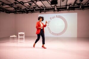 A presenter dressed in a red blazer, black cropped pants, and red shoes raises one finger as if making a point in front of a slide projected on a white wall with text displayed in a circle that reads For All Your Life. The word all has many l's in it, to drag the word on.