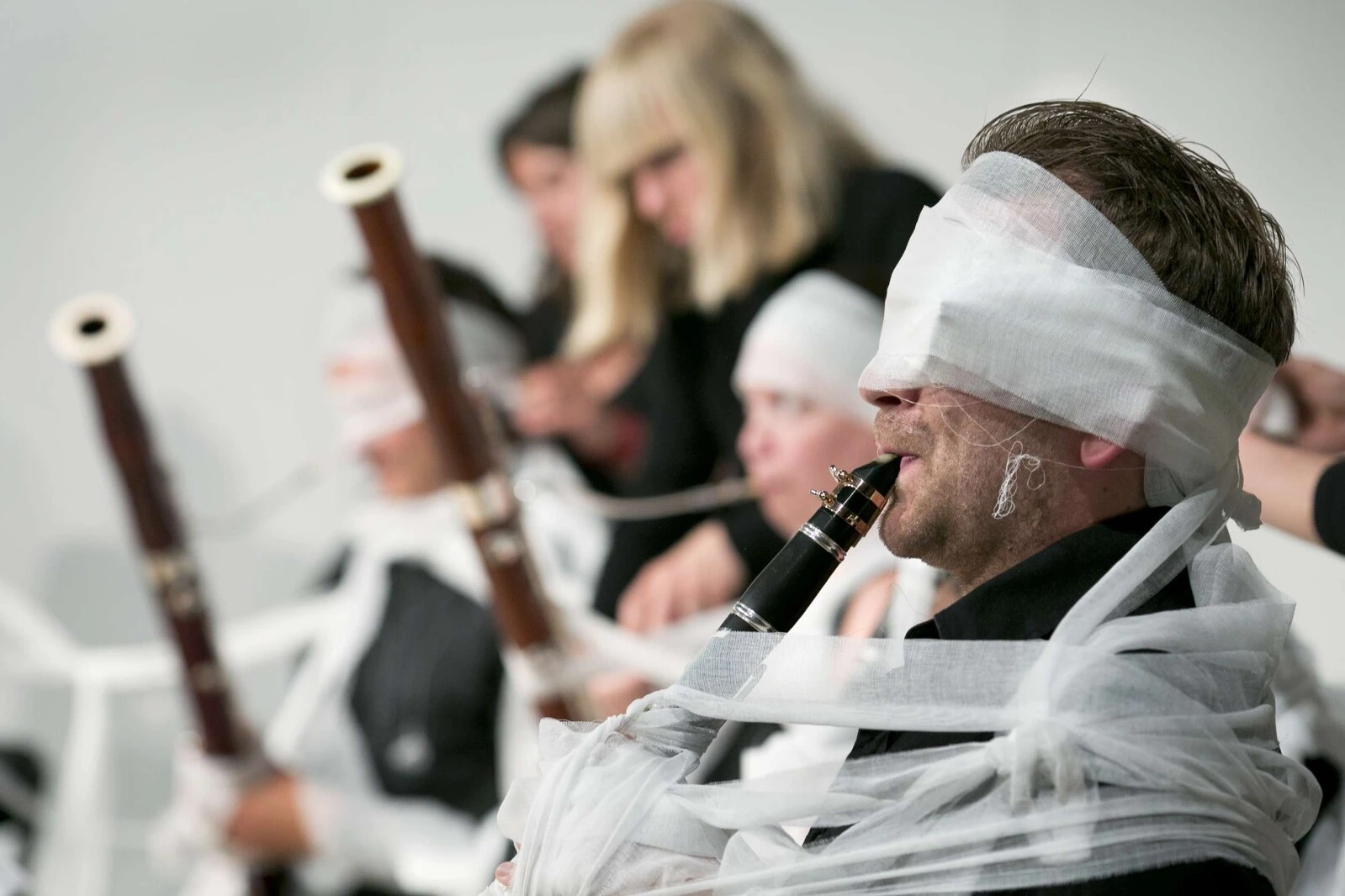 Musicians playing wooden recorders, seated in a row, wrapped in white gauze, some around their head as well.
