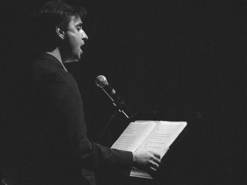 A male-presenting person reads from a stack of papers at a podium in a dark room