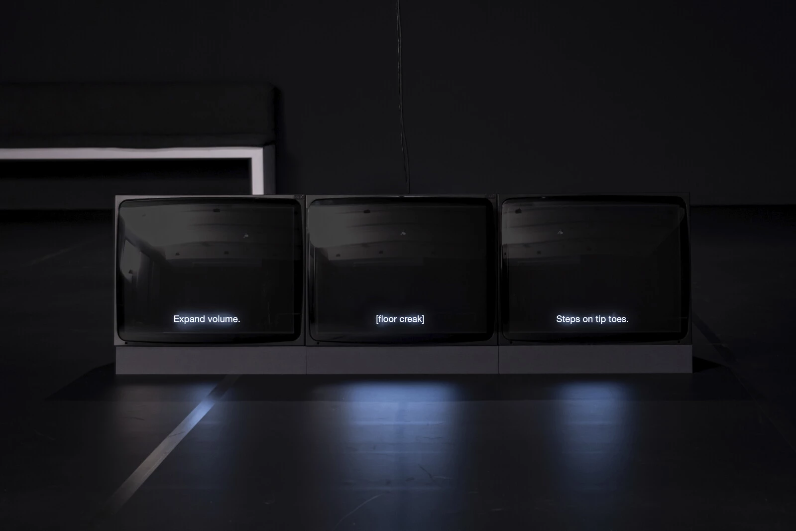 Three cube television monitors display monochrome black screens with white subtitles. From left to right, the subtitles read: