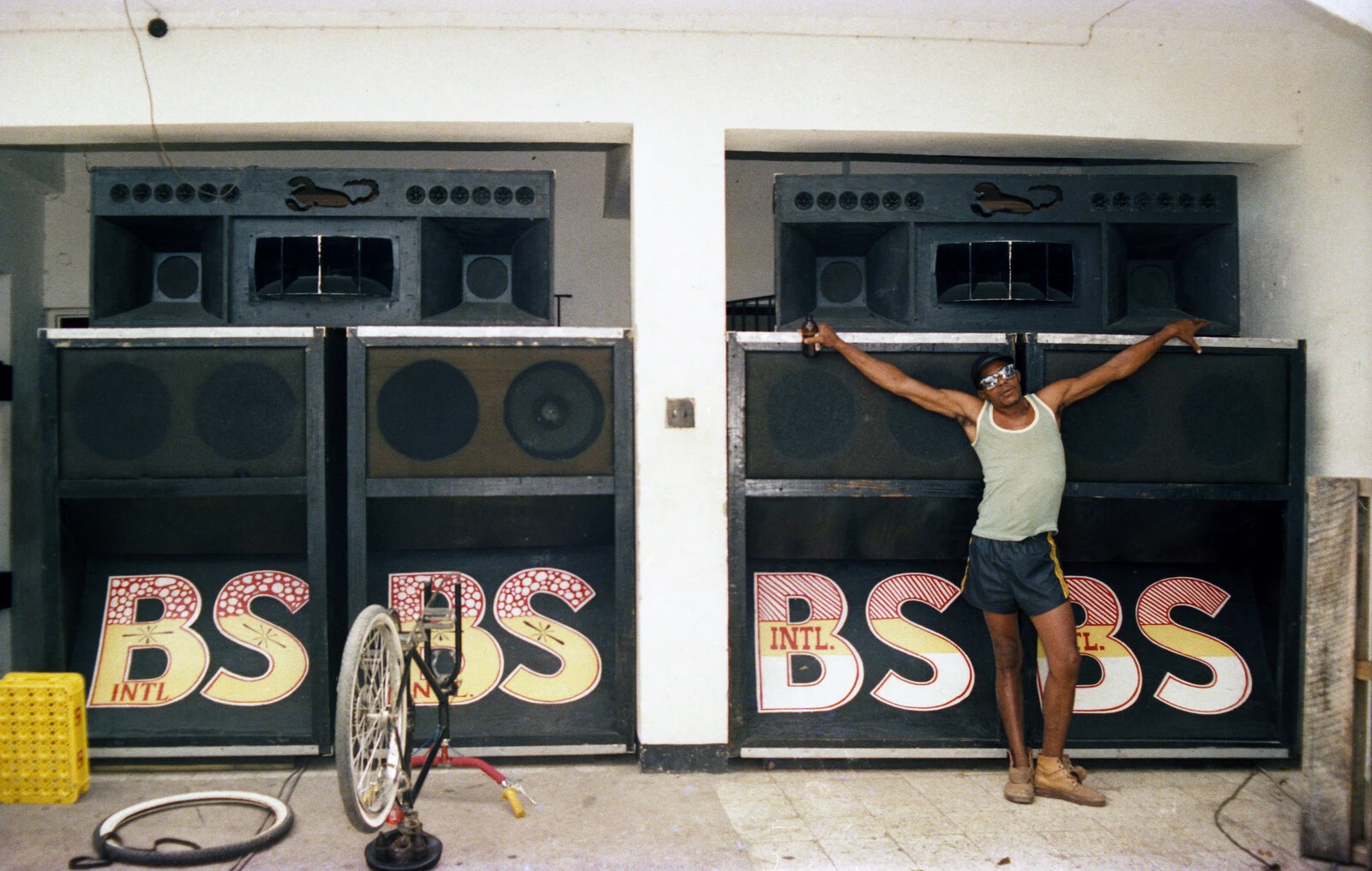 A person stands with arms stretched out, resting on two speaker systems that are taller than them in an alcove next to the same setup in an alcove to their right. Each has a logo of BS intl. on them.