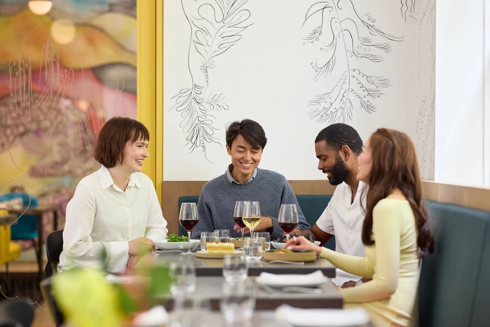 Four people sit at a round table in a brightly lit restaurant with floral line drawings on the walls