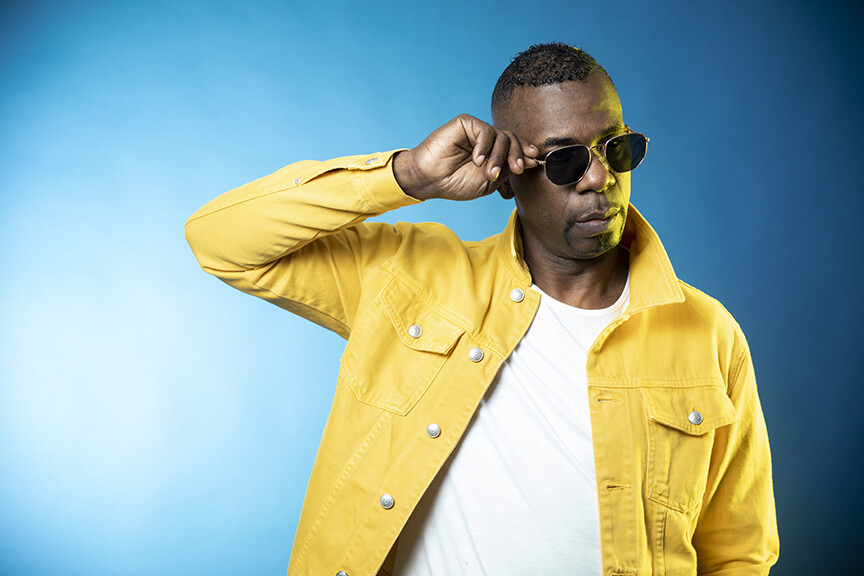 A person with dark skin wearing a bright yellow jacket and white t-shirt poses with their right hand to their sunglasses against a teal wall