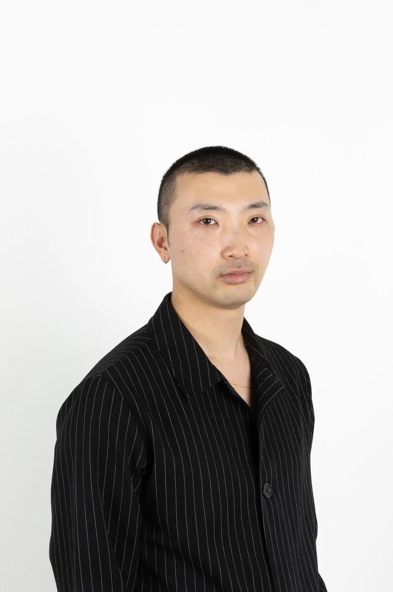 A male-presenting person with closely trimmed black hair and the shadow of a goatee poses against a bright white wall