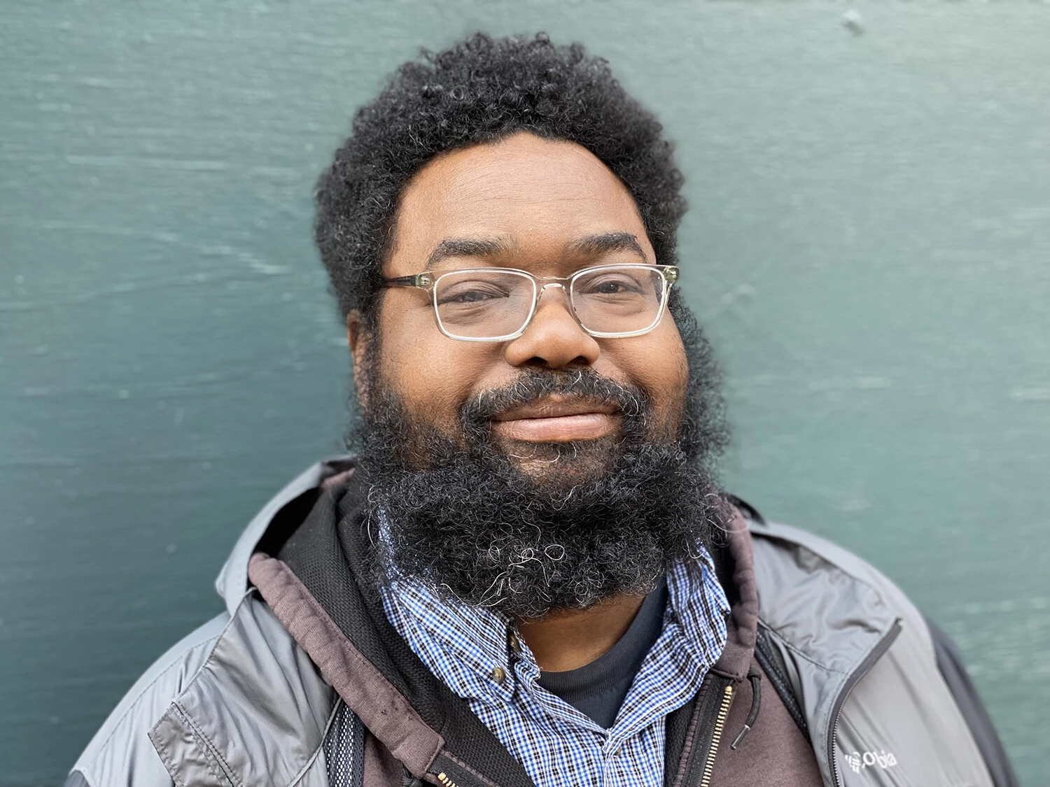 A male-presenting person in glasses with dark curly hair and a full beard poses against a teal wall