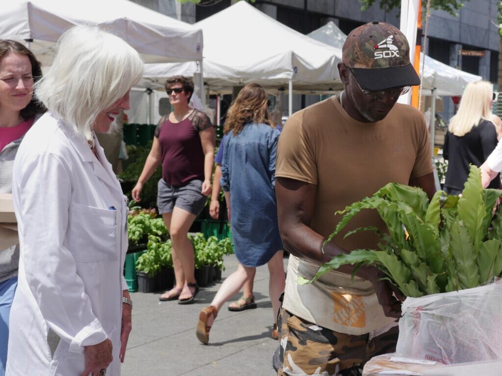 A man bags up leafy greens for another person outside with white tents and people walking by in the background